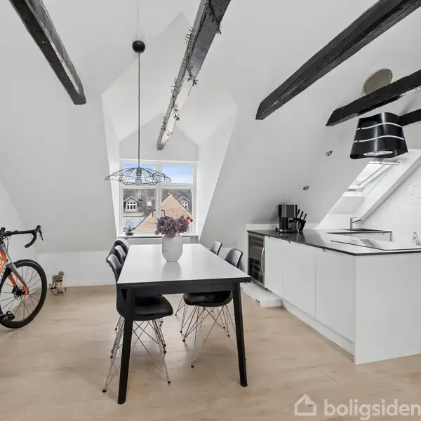 Cykel står op ad væg ved spisebord med fem sorte stole. Køkken med sort-hvide elementer ses til højre. Skrå lofter har sorte bjælker, og store vinduer giver udsigt over hustage.