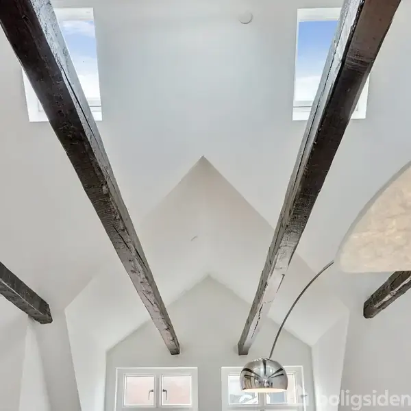 Mørke træbjælker strækker sig over en hvid loftsstruktur med trekantede vinduer, der lukker lys ind. En moderne lampe hænger fra loftet. Fotografier pryder den ene væg.