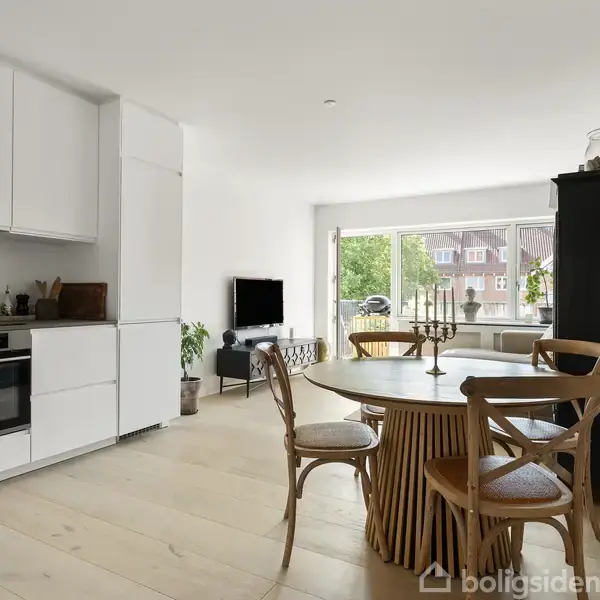 Et moderne køkken-alrum med hvide skabe og indbygget ovn, rundt spisebord med træstole. En stue med sofa, tv og stort vindue, der giver udsigt til grønne træer udenfor.