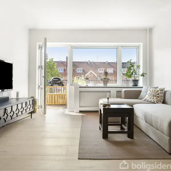 Stue med lys grå sofa, sofabord, og tv-bord med fladskærm. Glasdør fører til altan med udsigt over huse. Store vinduer lader lys ind, med planter på vindueskarmen.