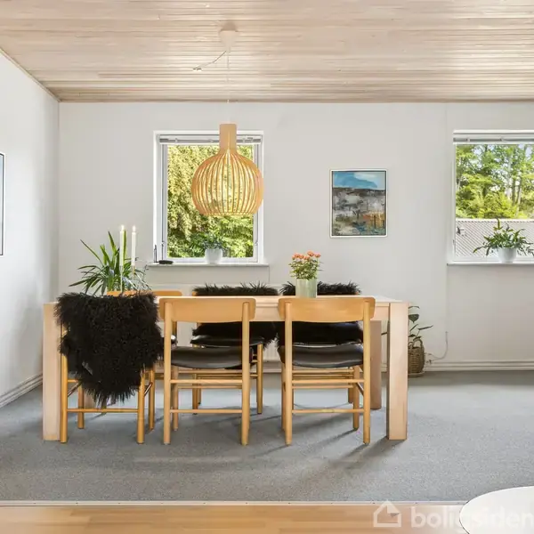 Spisebord med sorte stole står under en trælampe; et vindue med grøn udsigt til venstre, vægkunst til højre; en brændeovn står i højre hjørne af rummet.