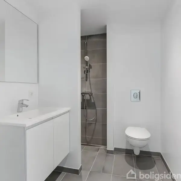 Badeværelse med håndvaskskab og spejl til venstre, toiletskål monteret på væg til højre, og bruseområde med grå fliser i baggrunden. Enkel og moderne indretning.