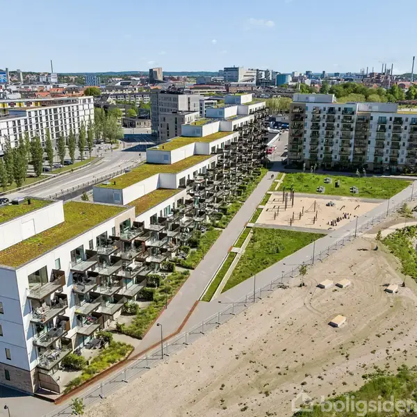 En række moderne lejlighedsbygninger med grønne tage strækker sig langs en vej. I forgrunden ses et åbent område med sand og en lille park med stier og græsplæner.