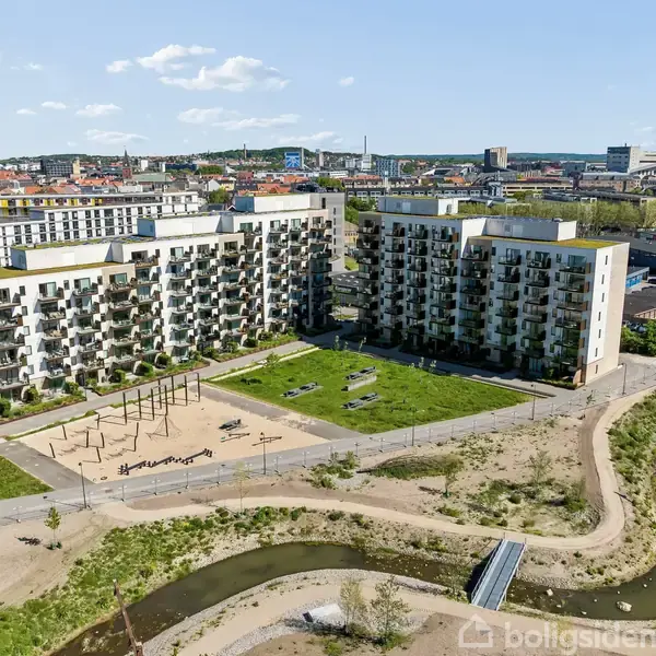 Lejlighedskompleks med altaner står ved en grøn park med legeplads. Små stier og en å løber gennem området. Bygninger og bymiljø ses i baggrunden.