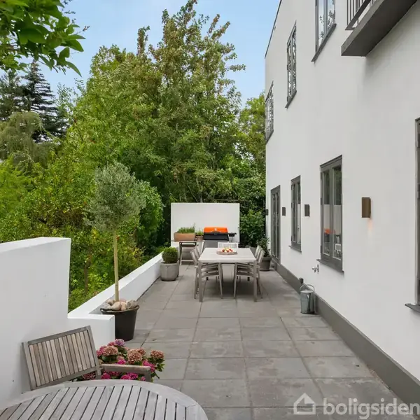 Terrasse med hvidmalet husvægge og grå fliser, indeholder et langbord med stole. Plantekrukker og blomster skaber en grøn atmosfære, omgivet af frodige træer. Ingen tekst til stede.