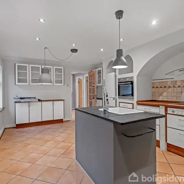 Et moderne køkken med en central køkkenø og integrerede apparater. Køkkenet har lyse vægge, flisegulv og hvide skabe med trædetaljer. Rummet er oplyst af indbyggede loftlamper.