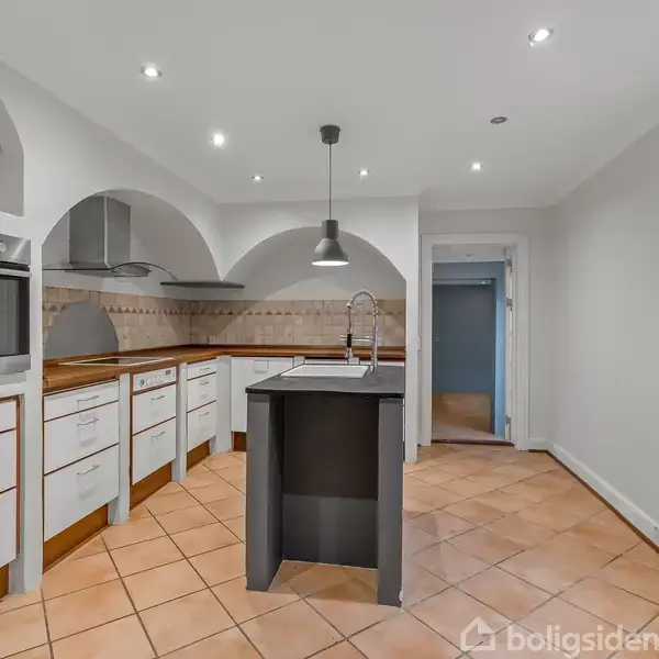 Et moderne køkken med lyse hvide skabe og en indbygget ovn. Der er en køkkenø med en vandhane i centrum, omgivet af lyse fliser og hvide vægge.