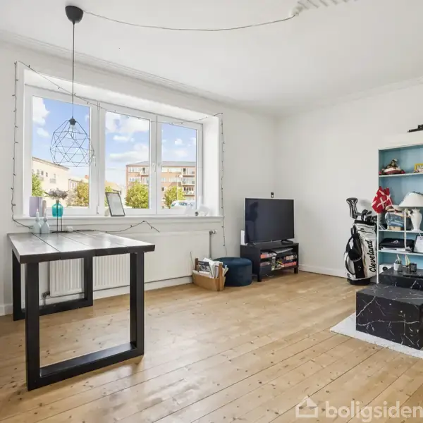 Stue med trægulv, stort vindue, spisebord og bænk. Reol fyldt med bøger og dekorationer, tv på lav skænk, golfsæt ved siden af. Ingen tekst er til stede.