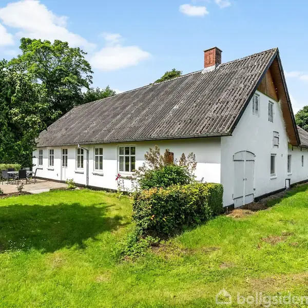 Hus med hvid facade og mørkt skråtag, stående på en grøn græsplæne. Omkranset af træer og buske under en blå himmel med få skyer.