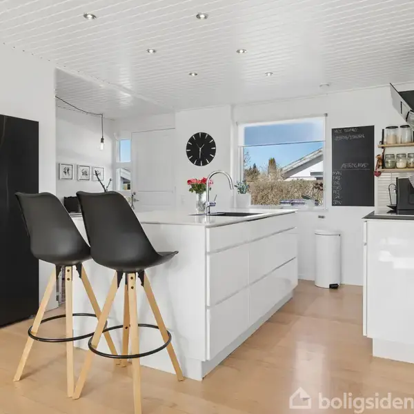 En moderne køkken med hvidt interiør domineret af en stor køkkenø. To sorte barstole står ved øen, og et stort sort køleskab er placeret til venstre. I baggrunden ses en opslagstavle.