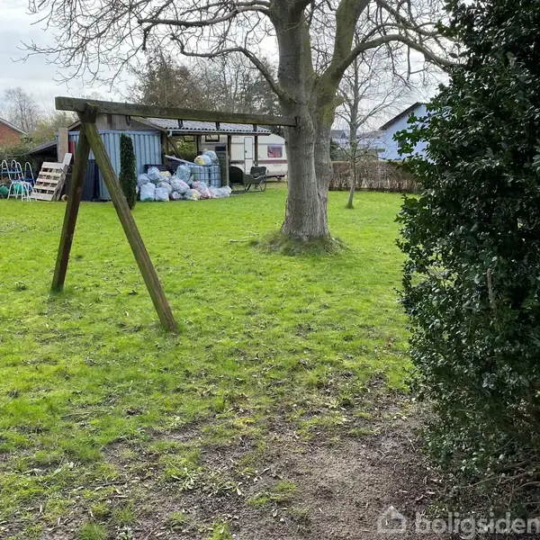 En tom trægyngestativ står på en grøn græsplæne ved siden af et stort træ. I baggrunden ses en skur fyldt med sække og en campingvogn.