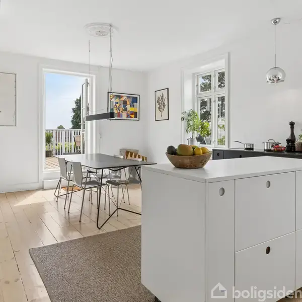 Et moderne køkken-alrum med et hvidt køkkenø, hvor en frugtskål står. Spisebord og stole står ved siden af en stor glasdør, med udsigt til en terrasse med grøn udsigt.
