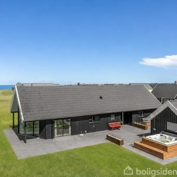 Sort, moderne hus med fladt tag står på græsplæne ved kysten. Der er store vinduer, en terrasse og en udendørs jacuzzi. Havudsigt i baggrunden under en klar blå himmel.