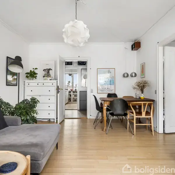 En moderne stue møbleret med en grå sofa og rundt sofabord, omgivet af lyse vægge og træmøbler. Et spisebord med sorte stole står ved siden af en kommode, planter og kunstneriske vægdekorationer.