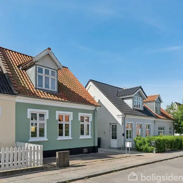 Række af charmerende huse med karnapper og farverige facader står langs en stille vej med en hvid stakit og grønne buske. Solrig himmel skaber en sommerlig atmosfære.