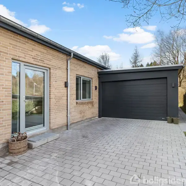 Hus i gule mursten med stor garageport, rottingkurv til venstre. Beliggende i en flisebelagt indkørsel omgivet af en velplejet have med træer og buske.