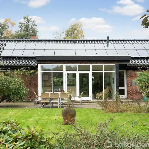 Et hus med solpaneler på taget står i en grøn have med græs og buske. Foran huset er der et terrasseområde med et bord og stole.