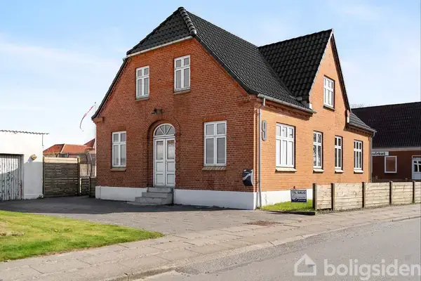 En rød murstenshus med sort tegltag står på en vej ved en fortov. Der er hvidmalede vinduer og en hoveddør med trapper. En til salg-skilt er ved siden af.