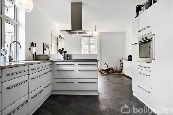 Et moderne køkken med hvide skabsfronter og indbygget ovn. En emhætte hænger over kogeøen, og store vinduer giver naturligt lys. Køkkenet har mørke gulvfliser og ligger i en lys, åben stue.