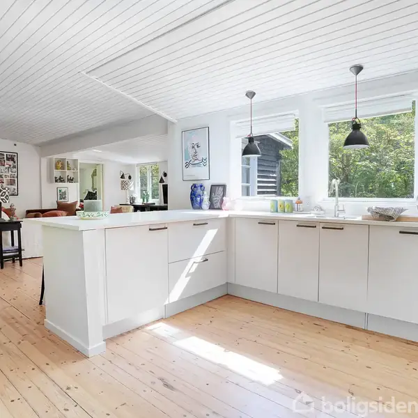 Et lyst køkken med lyse trægulve, hvide skabe og store vinduer, der slipper naturligt lys ind. Over kogeøen hænger to sorte lamper. I baggrunden ses dekorative elementer.
