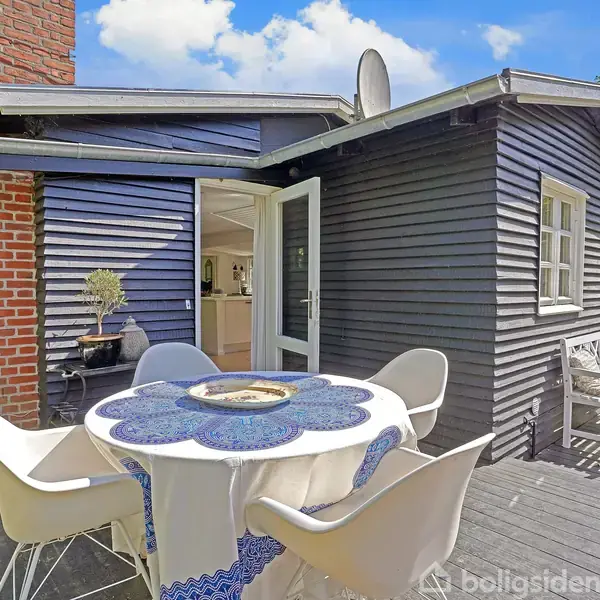 Et rundt bord med hvid dug og blå mønster er omkranset af fire hvide stole på en terrasse. Bagved ses et mørkeblåt træhus med en åben dør, omgivet af grøn have.