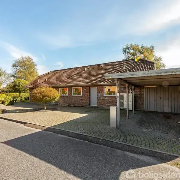 Et rødt murstenshus med brunt tag står stille ved siden af en carport. Det er omkranset af en flisebelagt indkørsel og vejen i et grønt beboelsesområde.