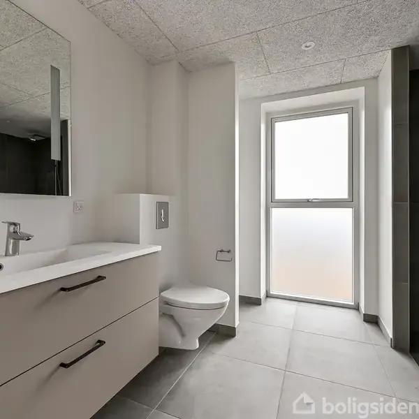 Badeværelse med håndvask og spejlskab til venstre, toilet i midten, og bruseniche til højre. Rummet har grå fliser på gulvet og et stort, frostet vindue i baggrunden.