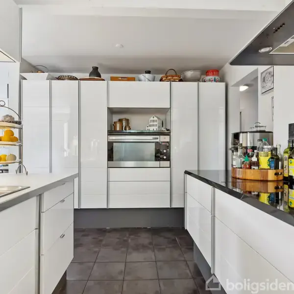 Et moderne køkken med hvide skabe og sort bordplade. Skabe rummer mikrobølgeovn og pynteting. På bordet står olieflasker og krydderiarrangement. Frugtkurv med citroner står ved siden af vasken.