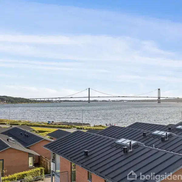 Flere huse står med mørke tage. I baggrunden ses en bro over en stor vandstrækning, omgivet af grønt landskab og en klar blå himmel.