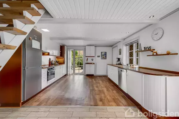 Køkken med trægulv og hvide skabe, indeholdende opvaskemaskine, ovn, og køleskab. Til højre en træhylde og ur på væggen. Glasdør åbner mod have. Ingen tekst fundet.