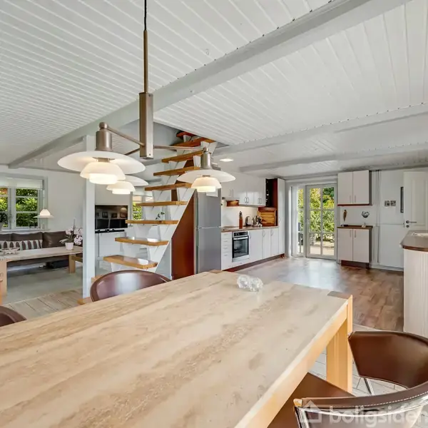 Et lyst køkken-alrum med træbord og stole står i forgrunden. En spiraltrappe leder til en ny etage. Rummet har åbne hylder, moderne apparater og store vinduer med udsigt.