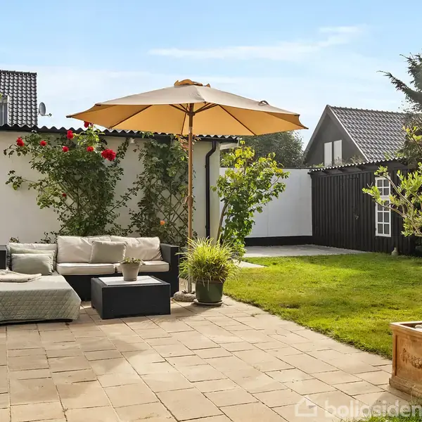 Terrassemøblement under en stor parasol står på flisebelagt område ved siden af grøn have med træer og buske. Et hvidt hus og et sort træskur ses i baggrunden.