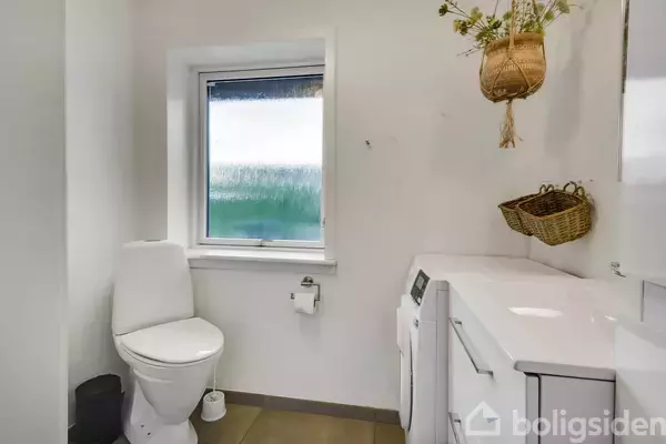 Toilet i et badeværelse ved siden af et vindue. Vaskemaskine og tørretumbler står til højre med kurve over sig. Rummet er lyst og enkelt indrettet.