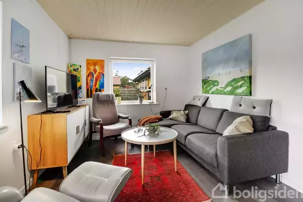 Grå sofa og lænestol står i stue med hvidt loft. Rundt sofabord på rødt tæppe. Tv på skænk, farverige malerier på væg, vindue ud til have.