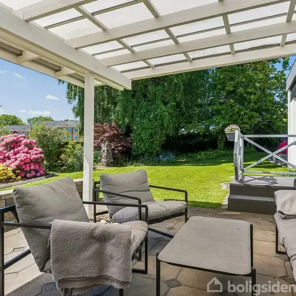 Terrasse med moderne møbler står under en hvid baldakin og tilbyder et hyggeligt opholdssted. I baggrunden ses en velholdt have med grønne træer og blomsterbede.