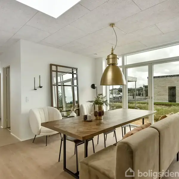 Et træbord står i en moderne spisestue med lyse møbler og en gylden lampe. Store vinduer viser en grøn udendørs scene med et fladt tagbyggeri i baggrunden.