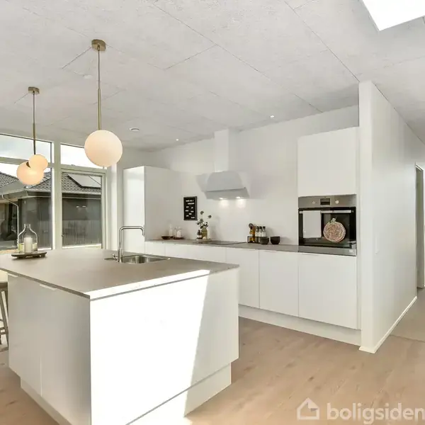 Et moderne køkken med en hvid køkkenø og indbygget ovn. Dagslys strømmer ind gennem store vinduer, der vender ud mod en græsplæne og en flisebelagt terrasse.