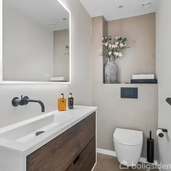 En moderne badeværelsesvask med mørke skuffer står stationær. En vase med blomster og toiletartikler pryder hylden. På væggen hænger et rektangulært spejl med belysning.