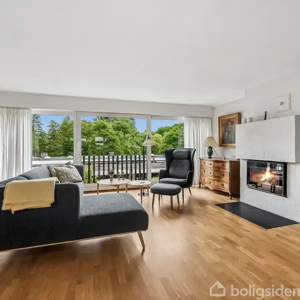 Sofa i moderne stue vender mod glasvæg, med udsigt til grøn beplantning udenfor. Komfortabel stol og pejs til højre. Trægulv og lyse vægge giver et let og åbent rum.