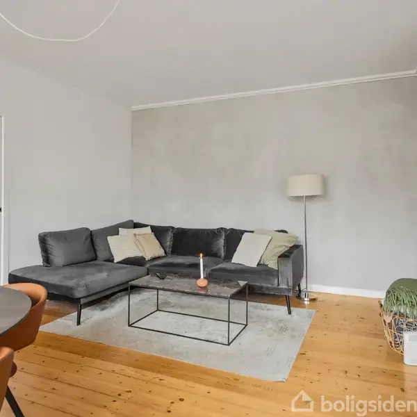 En grå hjørnesofa står på et lyst trægulv med et glassofa bord foran. En gulvlampe er i hjørnet, og et vægmonteret TV er til højre. Der står også en kurv på gulvet.