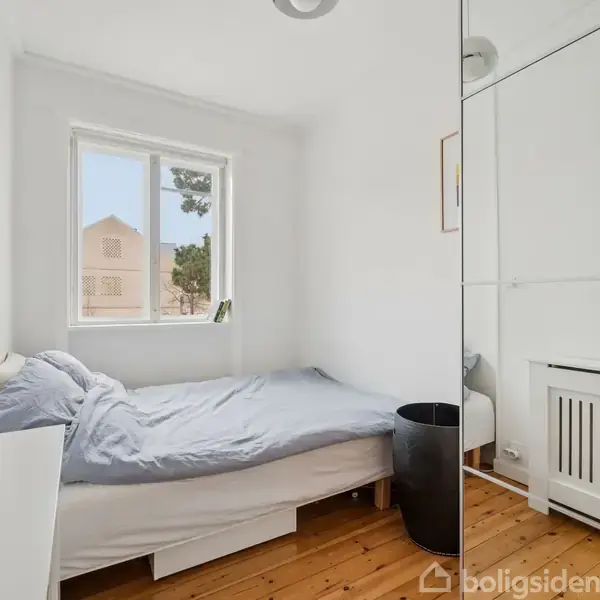 En seng står dækket med gråt sengetøj ved et vindue i et lyst rum. Trægulvet understøtter en moderne atmosfære; en skraldespand står ved siden af.