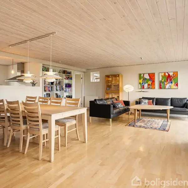 Træspisebord med otte stole står i en lys stue. To sofaer og et sofabord placeret på et tæppe. Væggene har farverige billeder, og der er store vinduer.