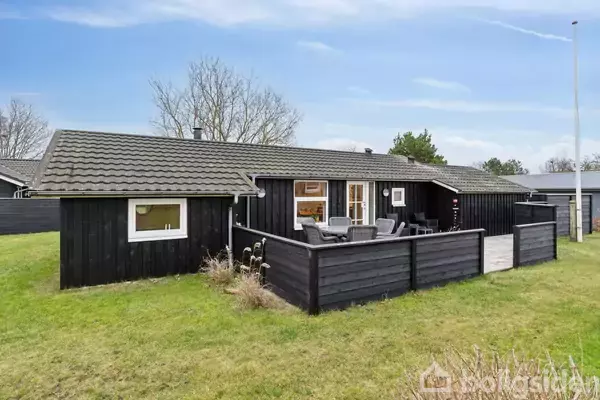Et sortmalet træhus med terrasse står på en velplejet græsplæne. Terrassen er møbleret med havestole og et bord. Omkringliggende træer skaber en rolig, naturlig atmosfære.