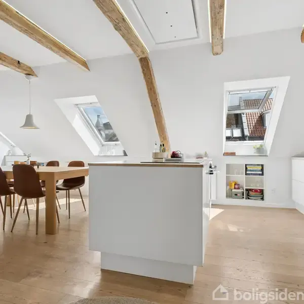 Et moderne køkken-alrum med træbjælker, hvor et spisebord med stole står ved siden af skråvinduer. Et køkkenø står centralt, og skuffer samt åbne hylder er langs væggen.