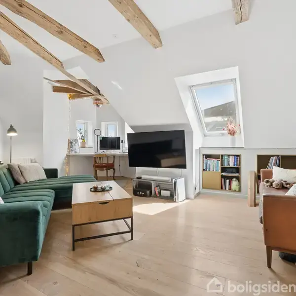 Stue med grøn sofa, TV på stativ og lænestol. Skråloft med synlige bjælker. Lysstrøm gennem ovenlysvindue. Hylder med bøger og dekorationer danner en hyggelig atmosfære.