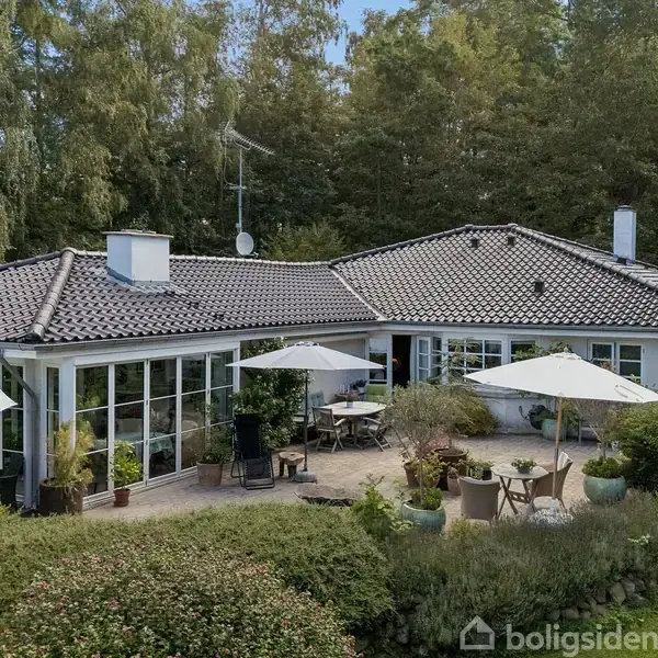 Hus i moderne stil står på en terrasse med havemøbler og hvide parasoller. Omgivet af grønne buske og træer i en skovlignende baggrund.