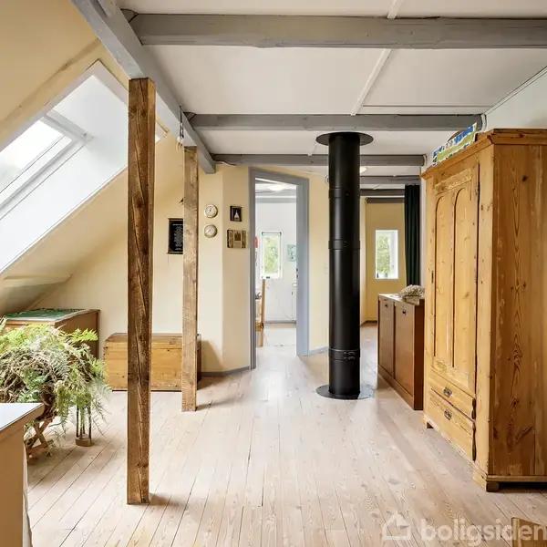 Et lyst loftsområde med naturligt lys fra skråvinduer. En sort brændeovn står centralt, omgivet af træmøbler og planter. Trælofter og -gulve skaber en hyggelig atmosfære.