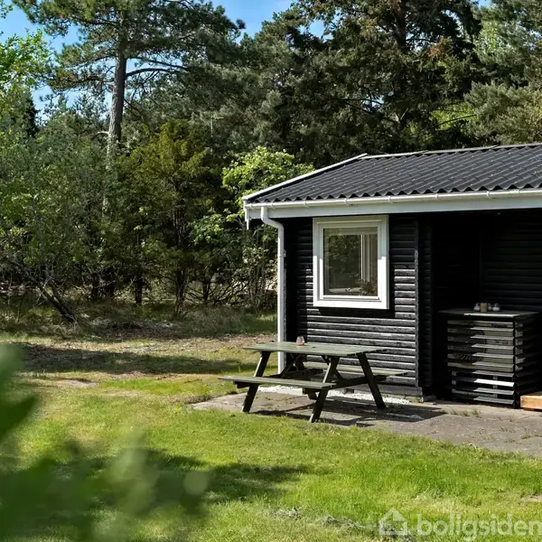 Sort træhus står stille i en grøn have med træer omkring. En træbænk står ved siden af huset, understreger sommerhusets afslappende atmosfære.