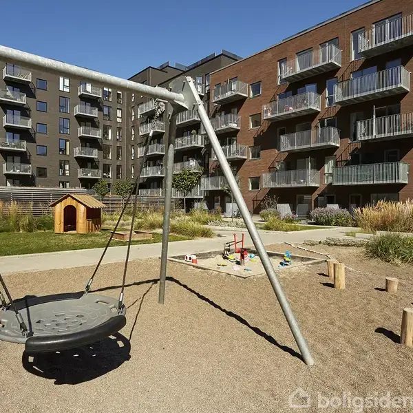 En sort gynge hænger i en ramme på en legeplads omgivet af sand. I baggrunden ses moderne lejlighedsbygninger og et lille legehus.