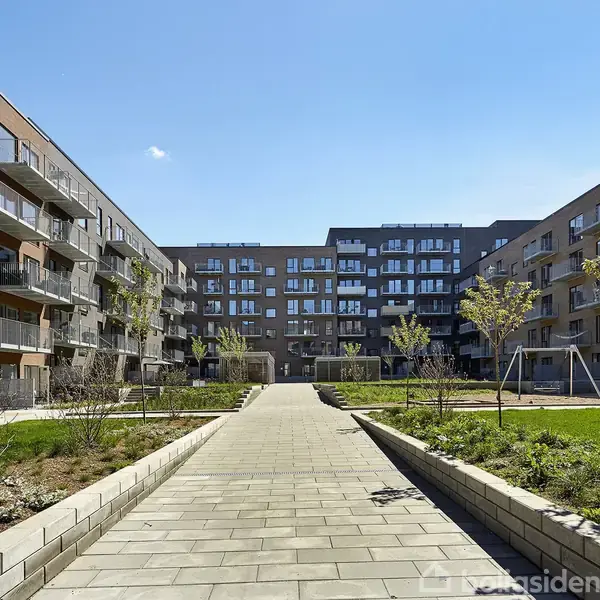 Et nybygget boligkompleks med altaner vender ud mod en åben gårdhave. Flisebelagt sti fører gennem området, omgivet af grønne områder og små træer under en klar, blå himmel.